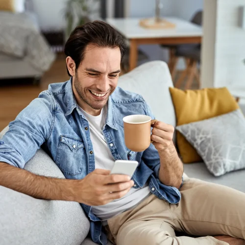 young-happy-man-texting-smart-phone-while-relaxing-sofa-drinking-coffee